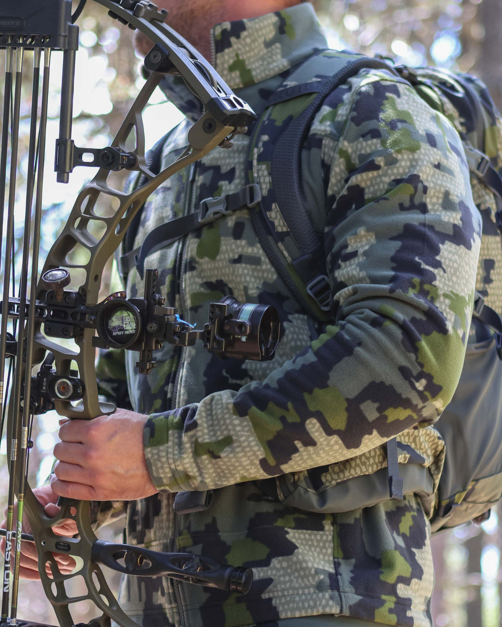 Man holds a bow upright next to his body wearing Border Fleece Full Zip Jacket in Verde Camouflage