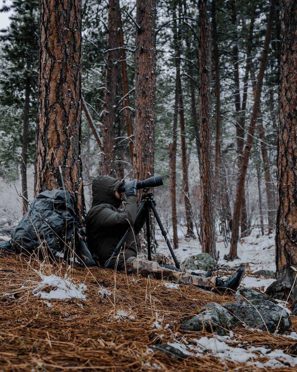 Side of Hunter Sitting Against Tree Glassing Game in Spotting Scope Wearing Super Down Burner Parka in Gunmetal Grey Color