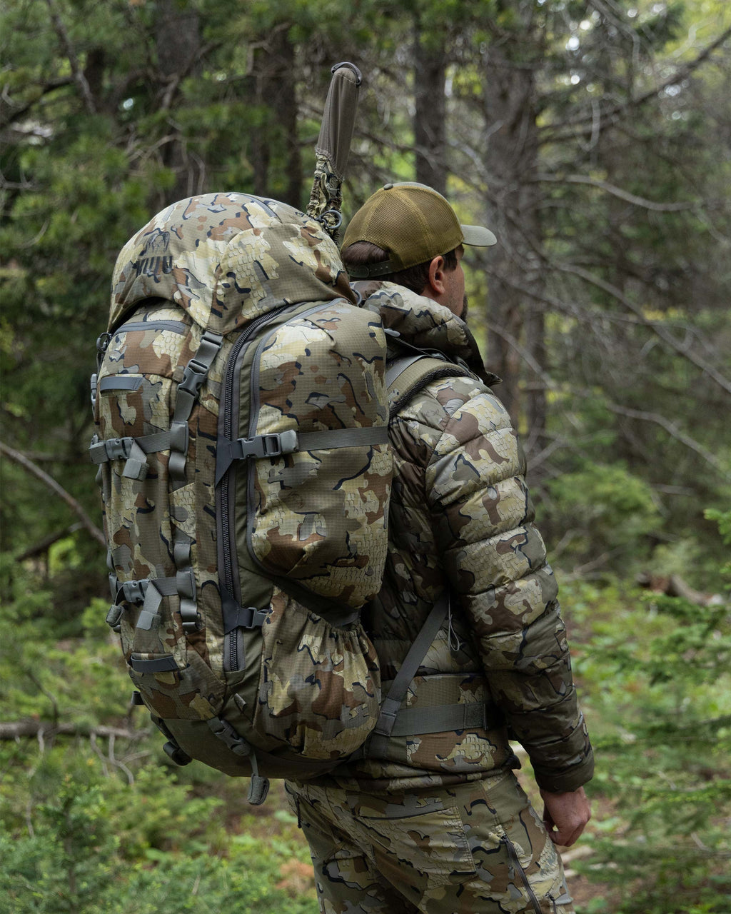 Hunter standing in a forest setting carrying an ICON 6 Divide 4500 Pack in Valo Camouflage with a covered rifle barrel protruding from the extended zipper opening.