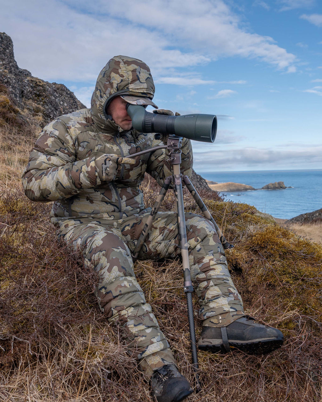 Hunter sitting in brush, glassing, wearing Super Down LT Hooded Jacket in Valo Camouflage