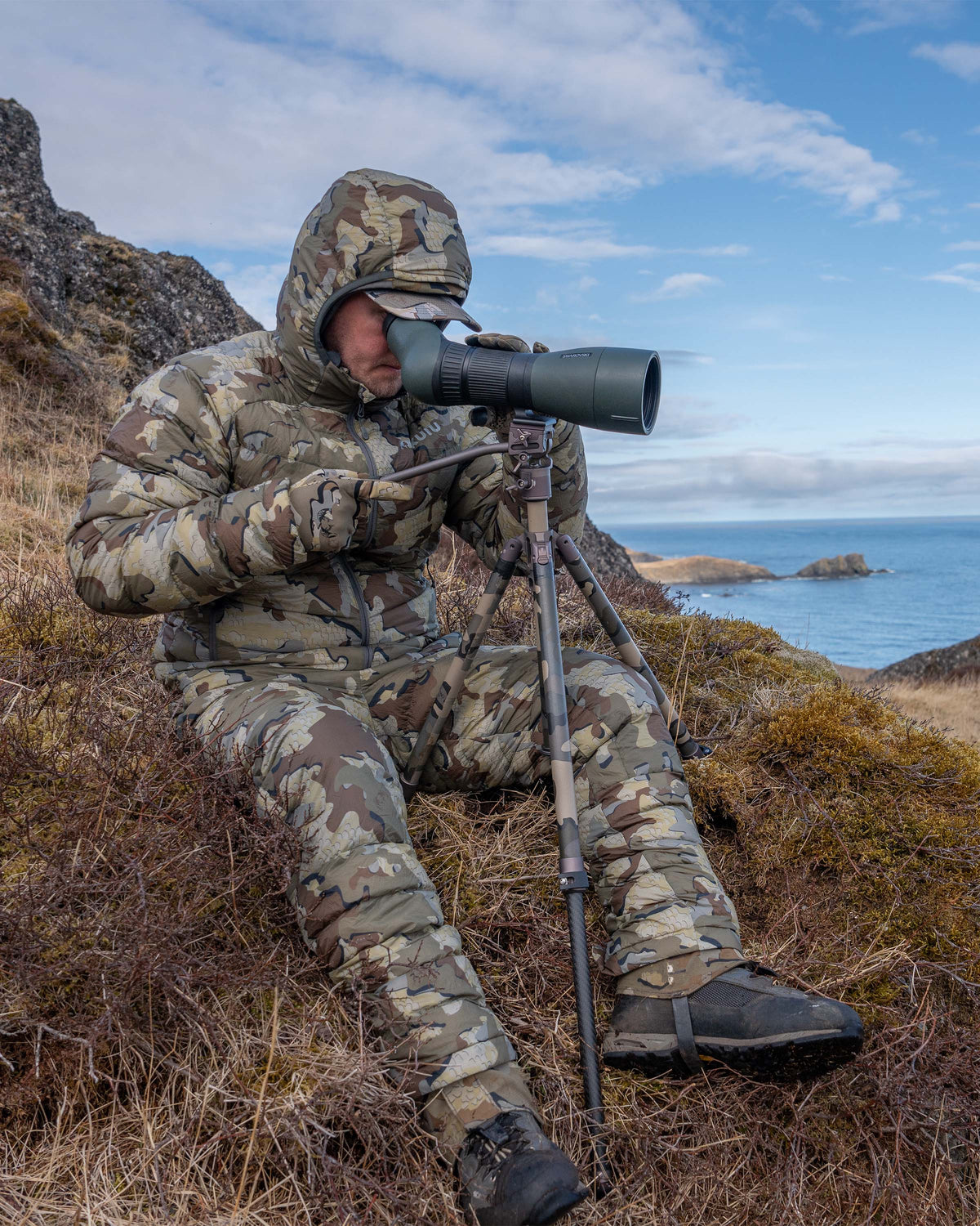 Hunter sitting in brush, glassing, wearing Super Down LT Hooded Jacket in Valo Camouflage