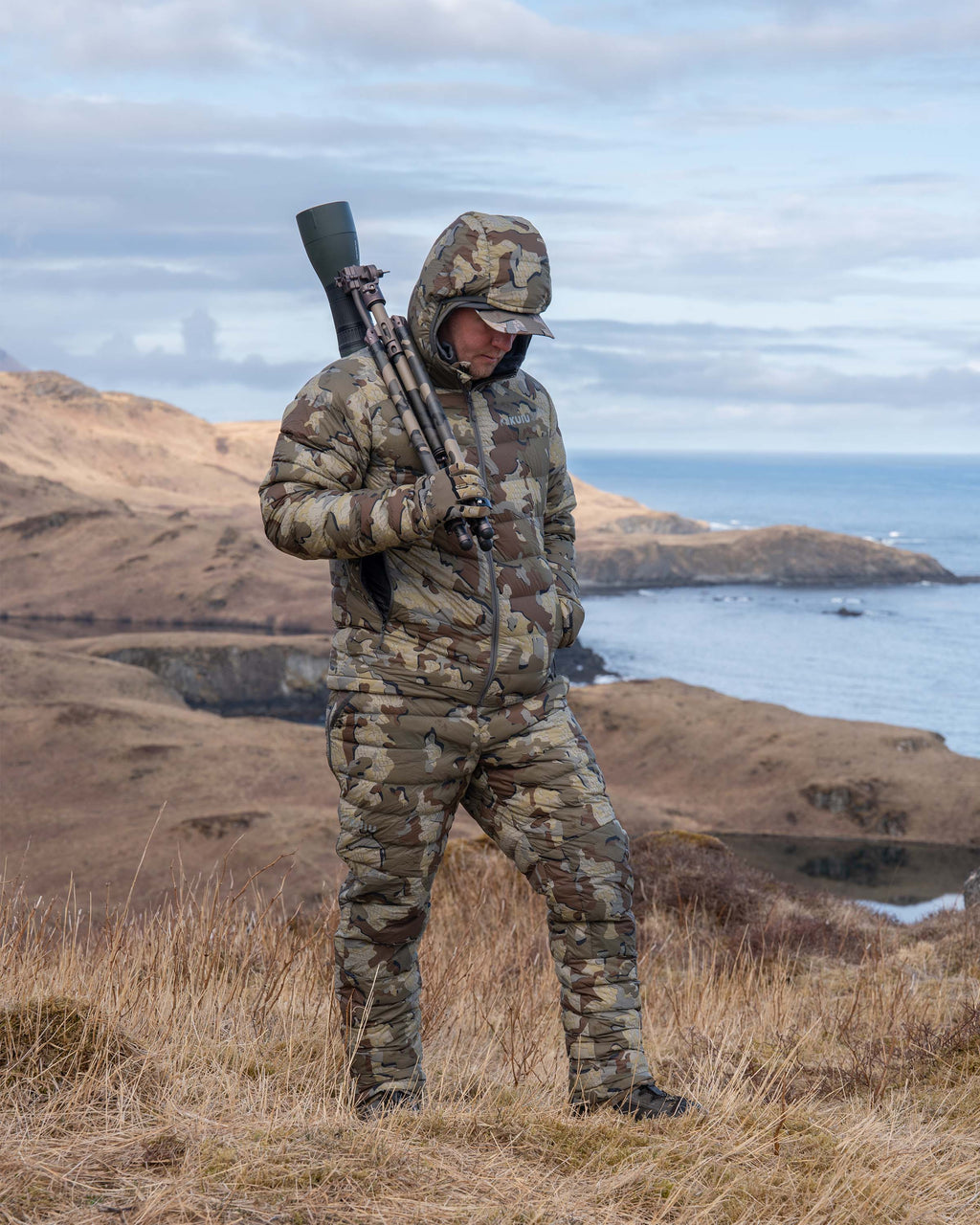 Hunter walking with glassing tripod wearing Super Down LT Hooded Jacket in Valo Camouflage