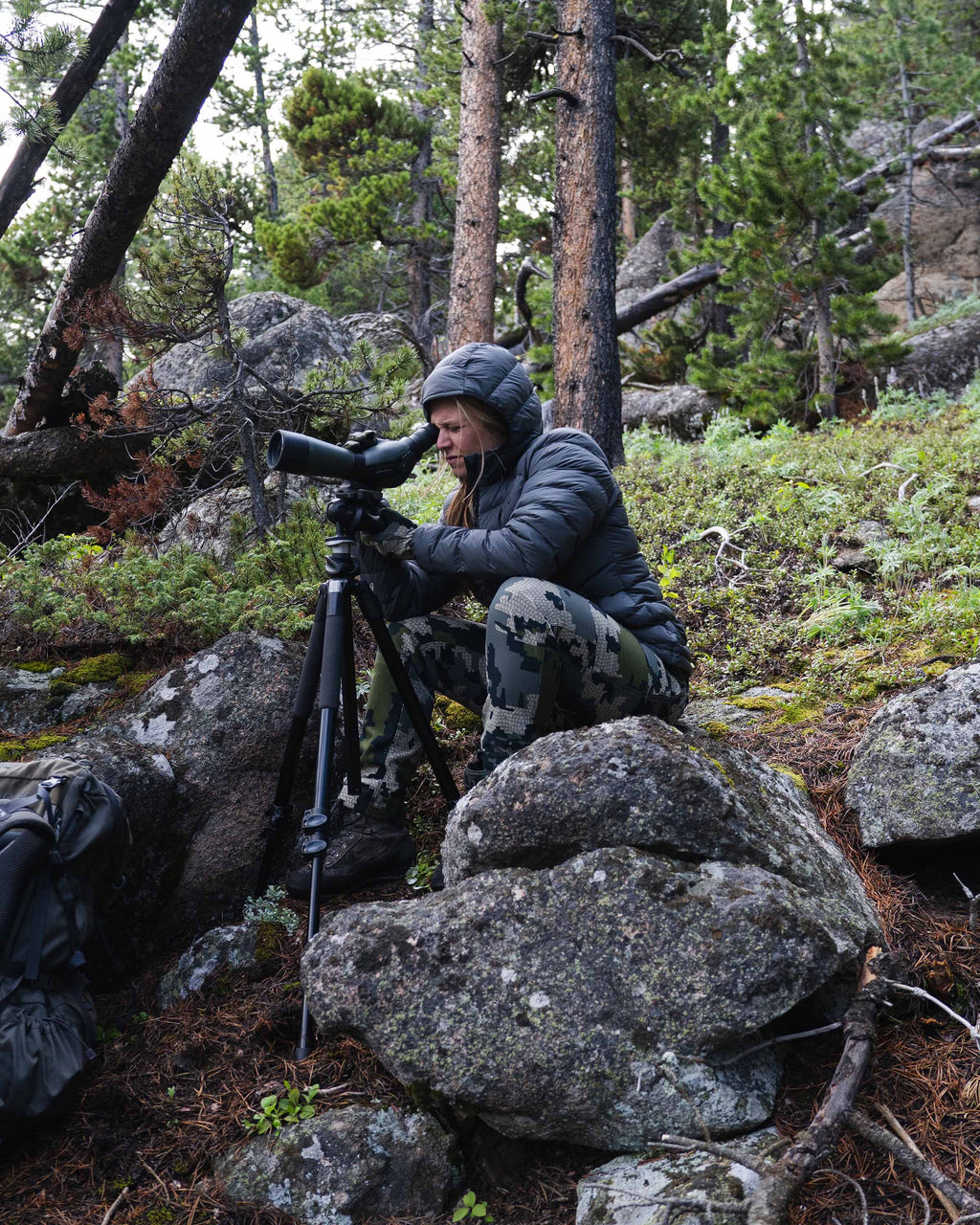 Hunter Glassing Game Through Spotting Scope on Wooded Hillside Wearing Women's Super Down LT Hooded Jacket in Black Color