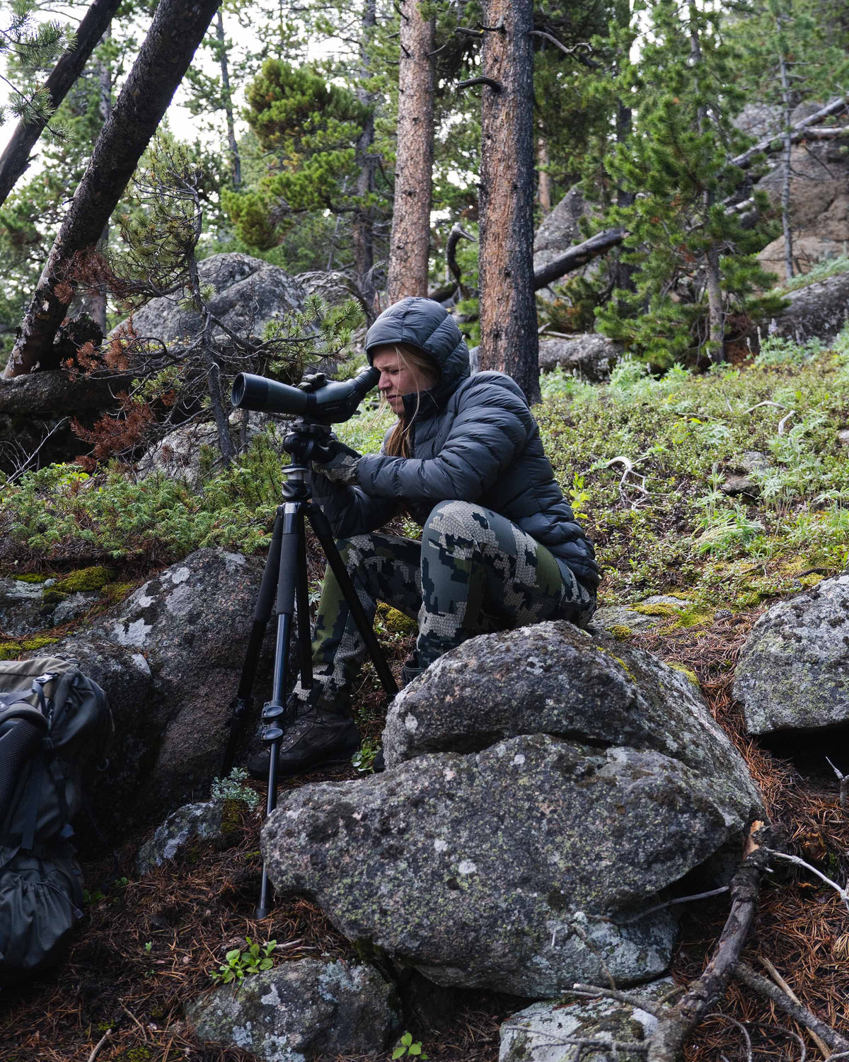 Hunter Glassing Game Through Spotting Scope on Wooded Hillside Wearing Women's Super Down LT Hooded Jacket in Black Color