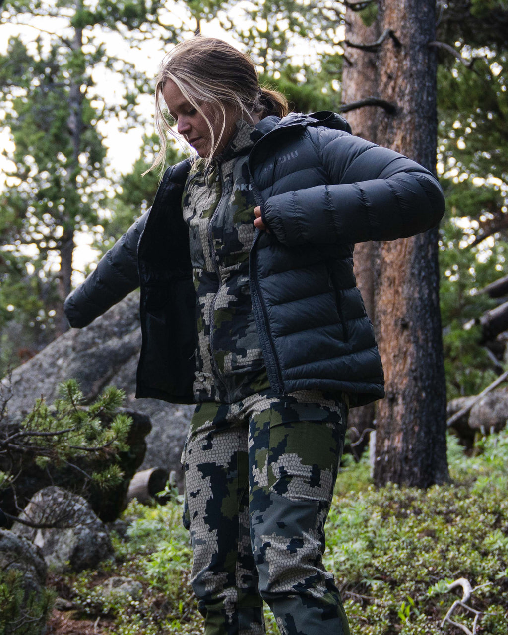 Hunter Standing in Wooded Terrain Putting on a Women's Super Down LT Hooded Jacket in Black Color