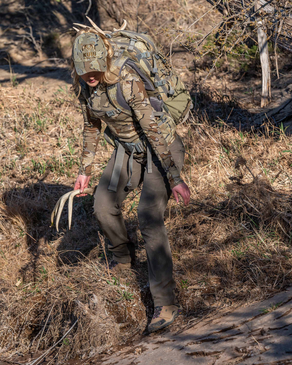 Angled, Front View of Hunter Holding Shed While Standing in Dry Grass Wearing Women's Attack Pants in Ash Brown Color
