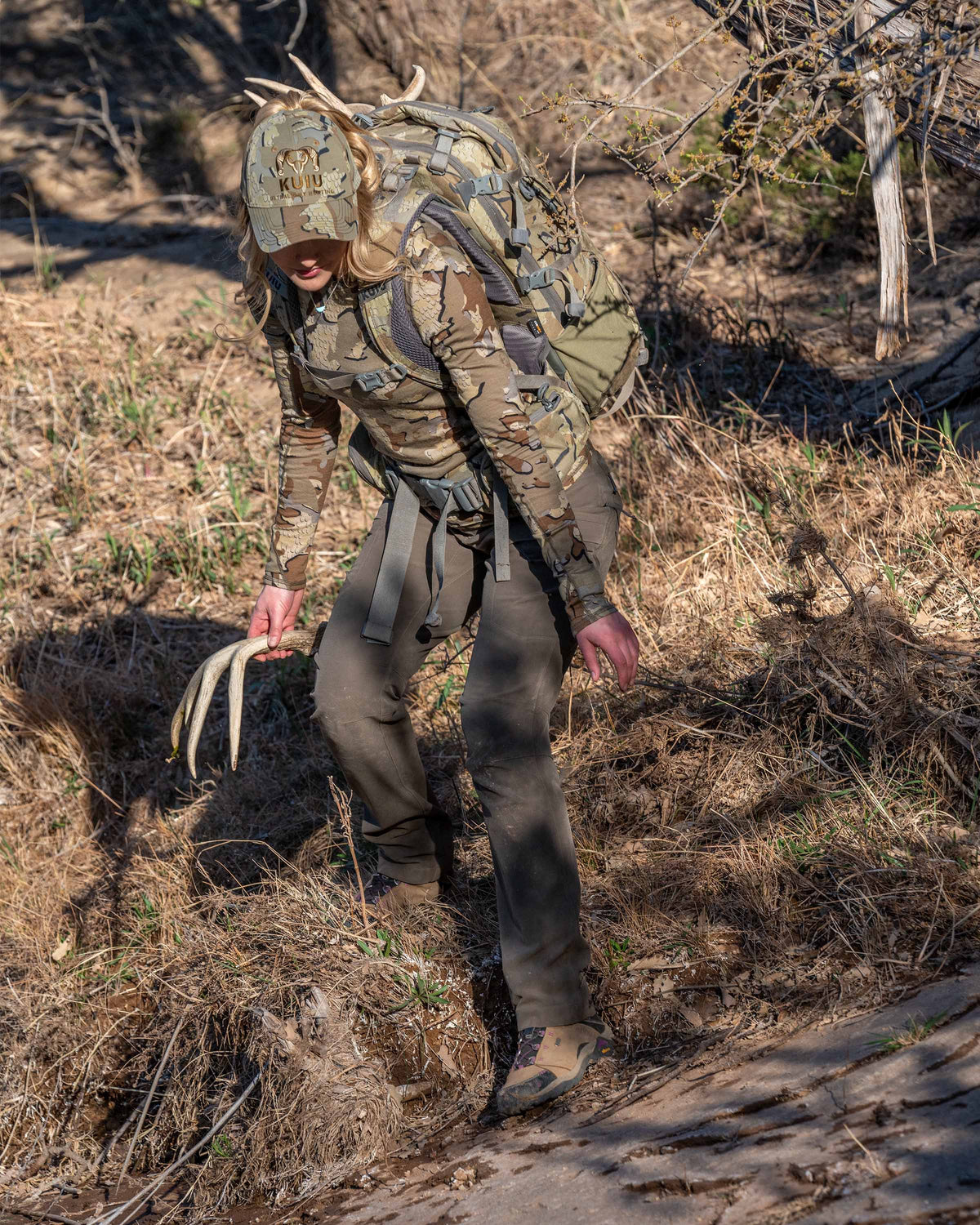 Angled, Front View of Hunter Holding Shed While Standing in Dry Grass Wearing Women's Attack Pants in Ash Brown Color