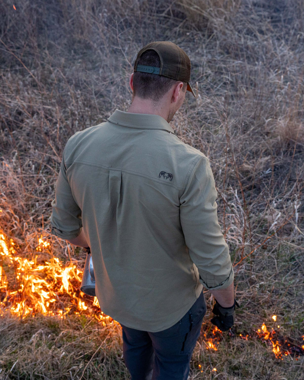 Back view of Hunter in a grass field, backburning using a drip torch, wearing Motive LS Work Shirt in Khaki