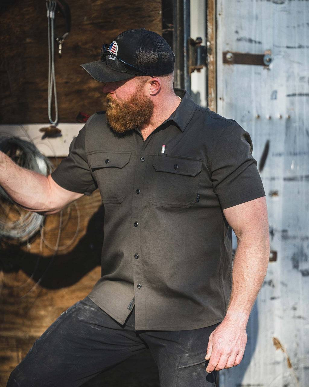 Man wearing Motive Short Sleeve Workgrade Shirt in Asphalt Grey standing in a workshop leaning against a metal sliding door.