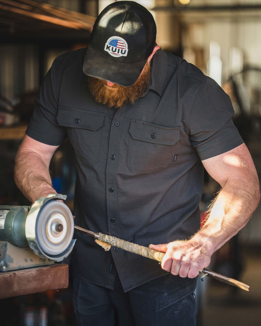 Man wearing Motive Short Sleeve in Asphalt Grey operating a bench grinder to shape a metal rod in an indoor workshop.