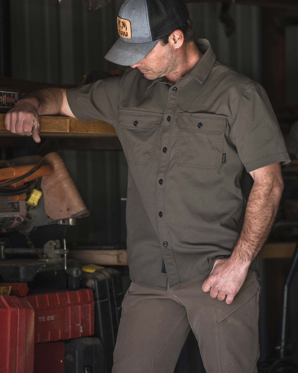 Man wearing a Motive Short Sleeve Workgrade Shirt in Rodeo Brown standing in a workshop and leaning an arm on a wooden shelf beside power tools and storage cases.
