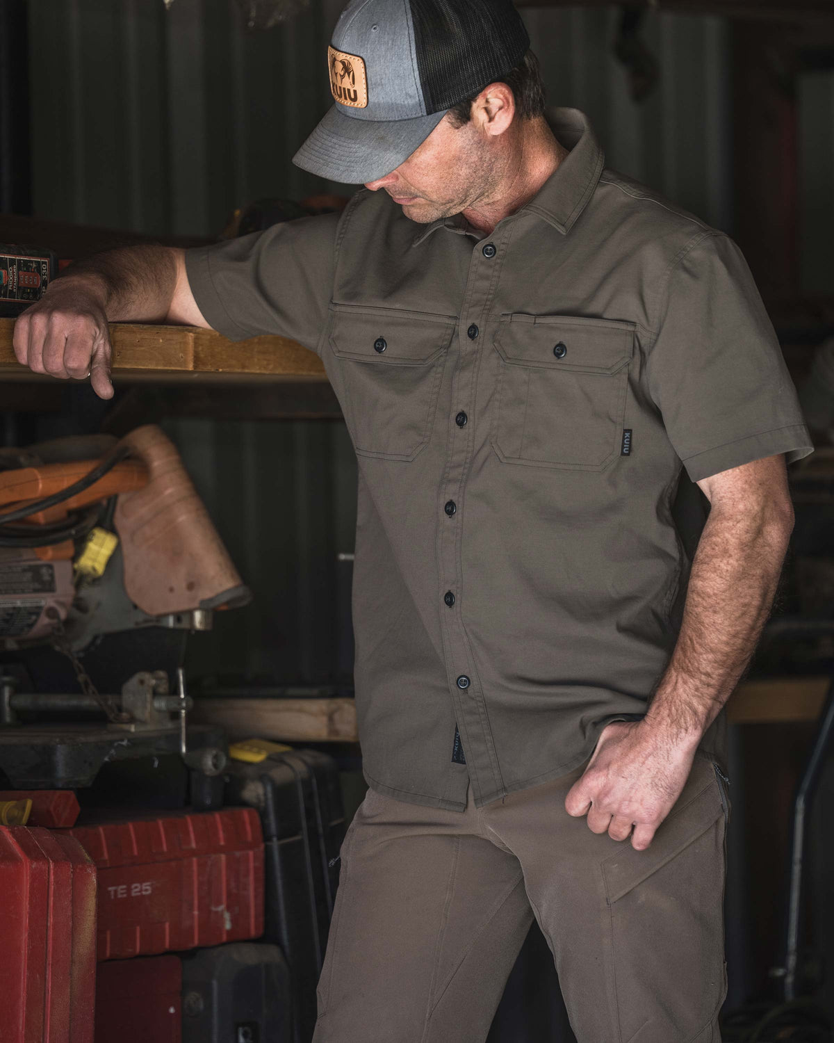 Man wearing a Motive Short Sleeve Workgrade Shirt in Rodeo Brown standing in a workshop and leaning an arm on a wooden shelf beside power tools and storage cases.
