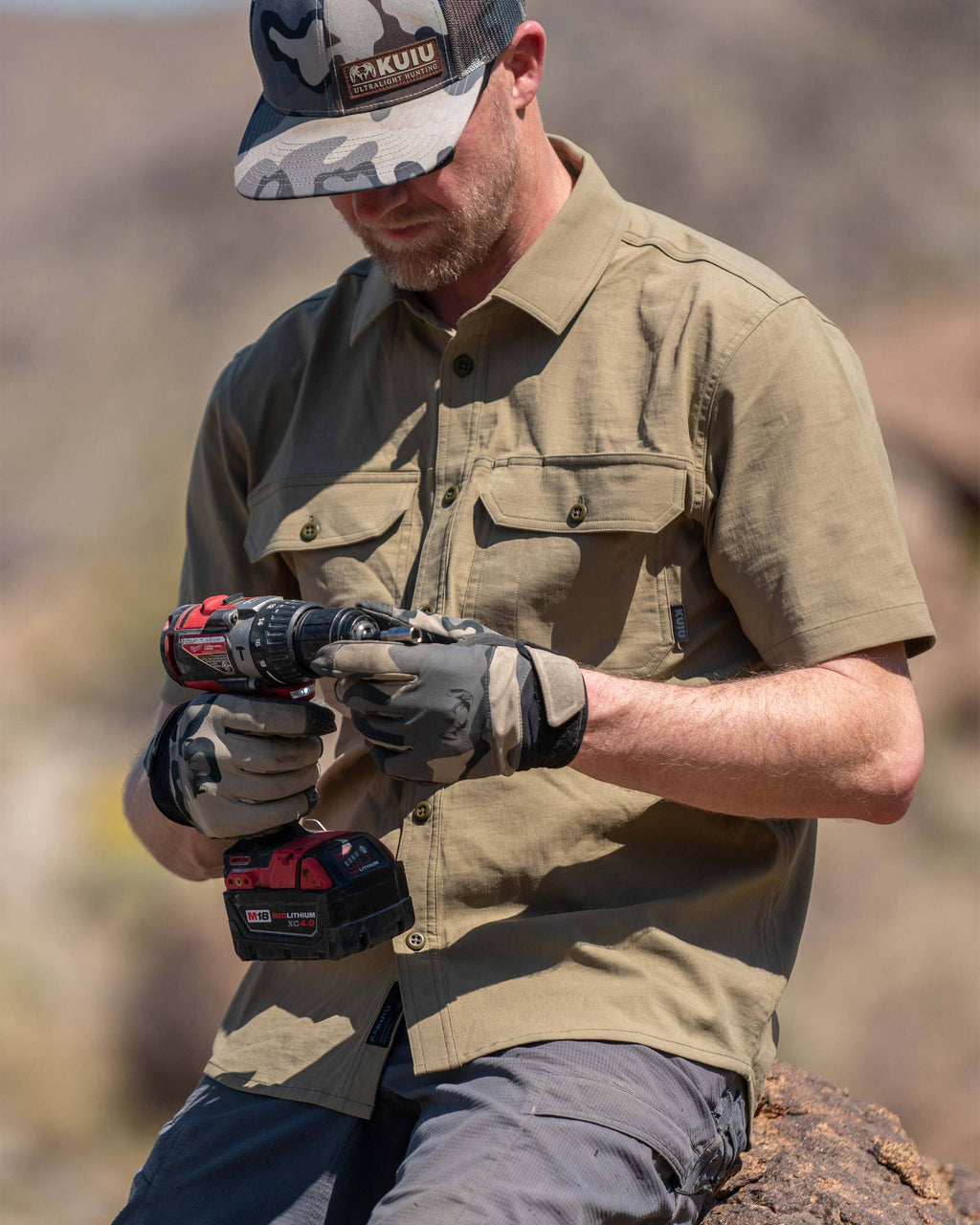 Model sitting on a rock, looking at his cordless screwdriver, wearing Motive Short Sleeve Work Shirt in Khaki