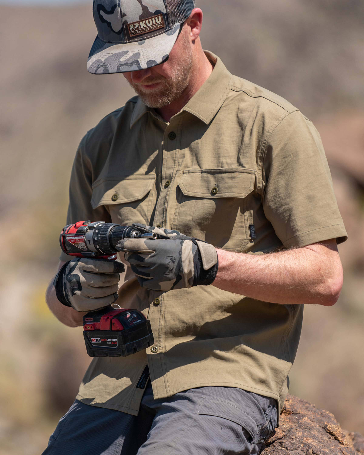 Model sitting on a rock, looking at his cordless screwdriver, wearing Motive Short Sleeve Work Shirt in Khaki