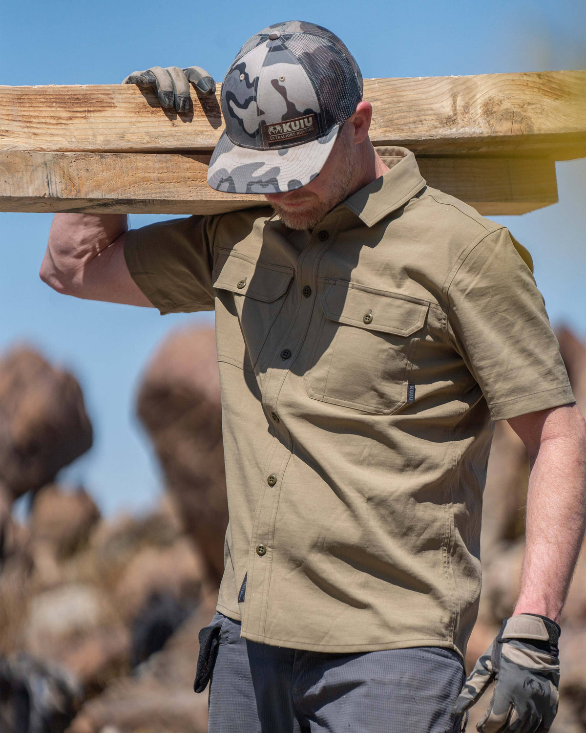 Model carrying three pieces of lumber on his right shoulder, wearing Motive Short Sleeve Work Shirt in Khaki