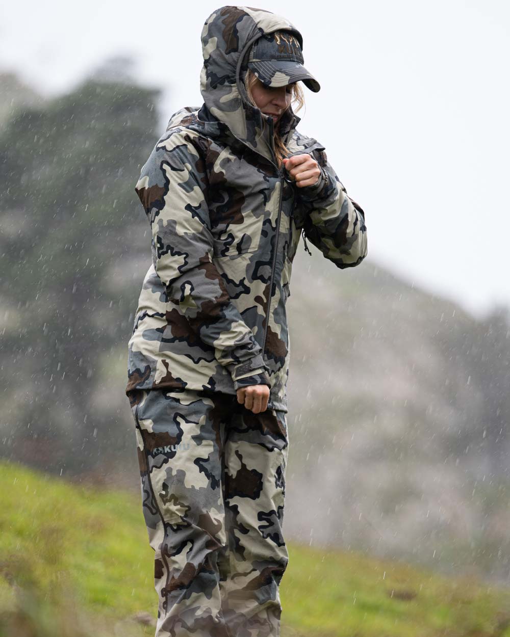 Angled, Front View of Hunter Standing in the Rain Zipping Up Women's Chugach TR Rain Jacket in Vias Camouflage