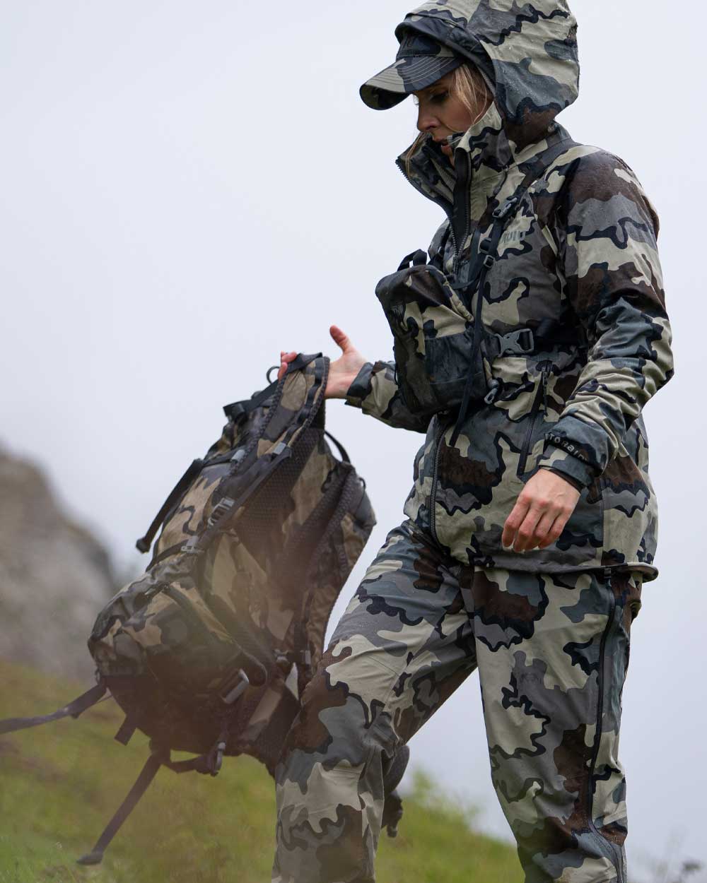 Hunter Holding Backpack Wearing Women's Chugach TR Rain Jacket in Vias Camouflage