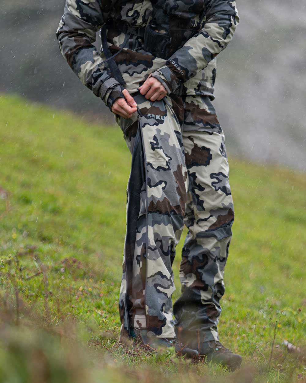 Hunter Standing in the Rain on Green Hillside Zipping Up Full Length YKK Leg Zippers on Women's Chugach TR Rain Pants in Vias Camouflage