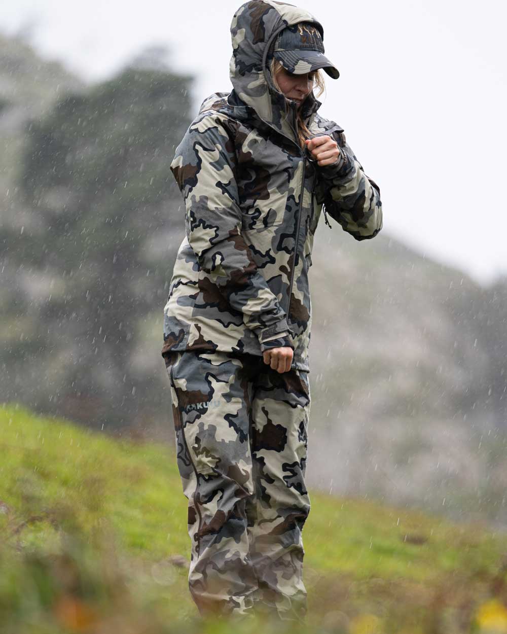 Hunter Standing in the Rain on Green Hillside Wearing Women's Chugach TR Rain Pants in Vias Camouflage
