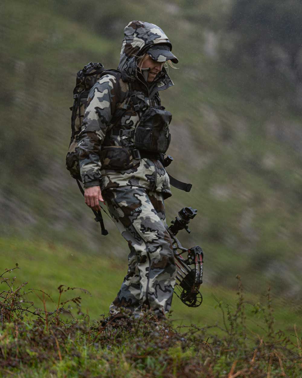 Hunter Walking in the Rain on Green Hillside Wearing Women's Chugach TR Rain Pants in Vias Camouflage