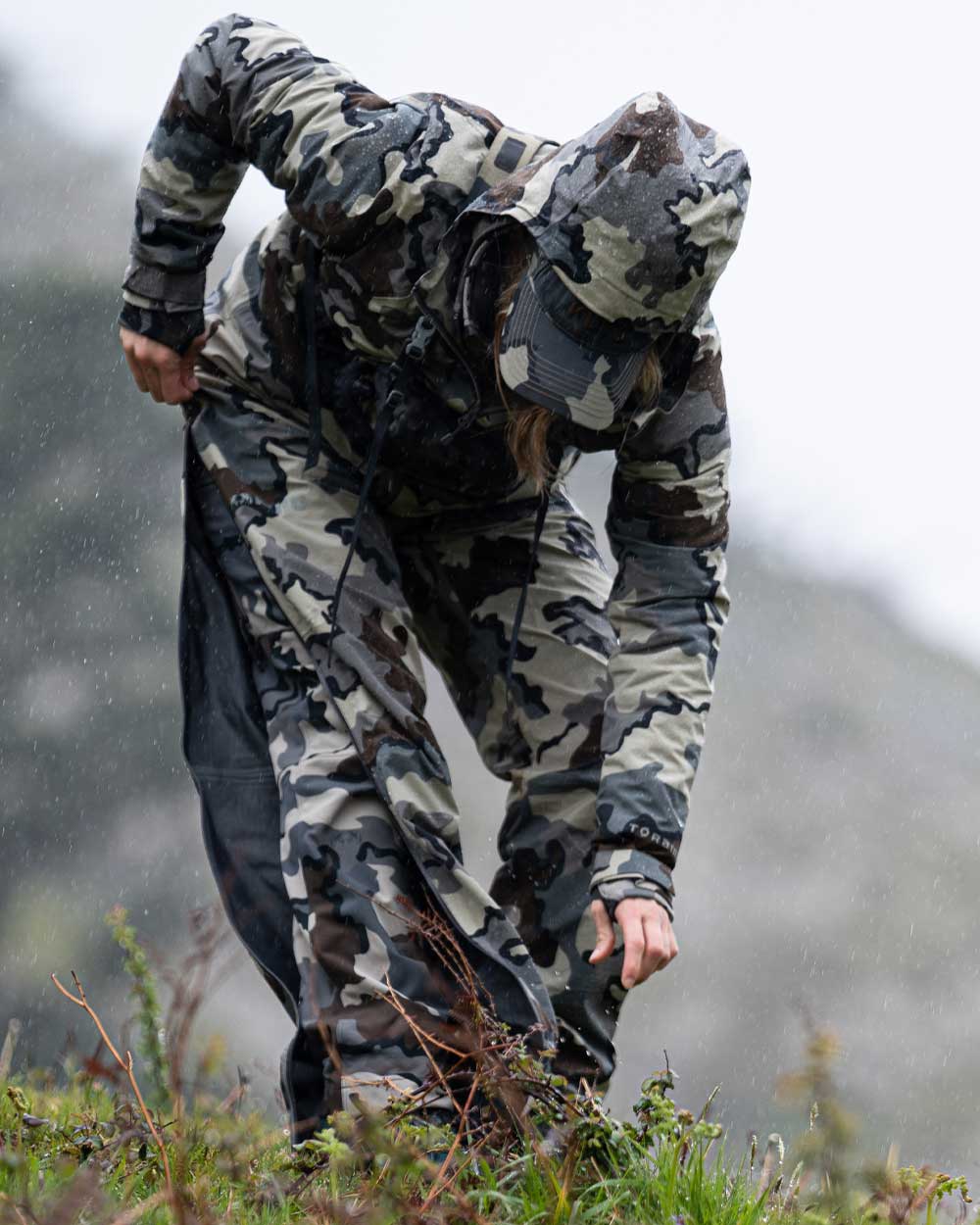 Hunter Standing in the Rain Putting on Women's Chugach TR Rain Pants in Vias Camouflage Highlighting Full Length YKK Leg Zippers