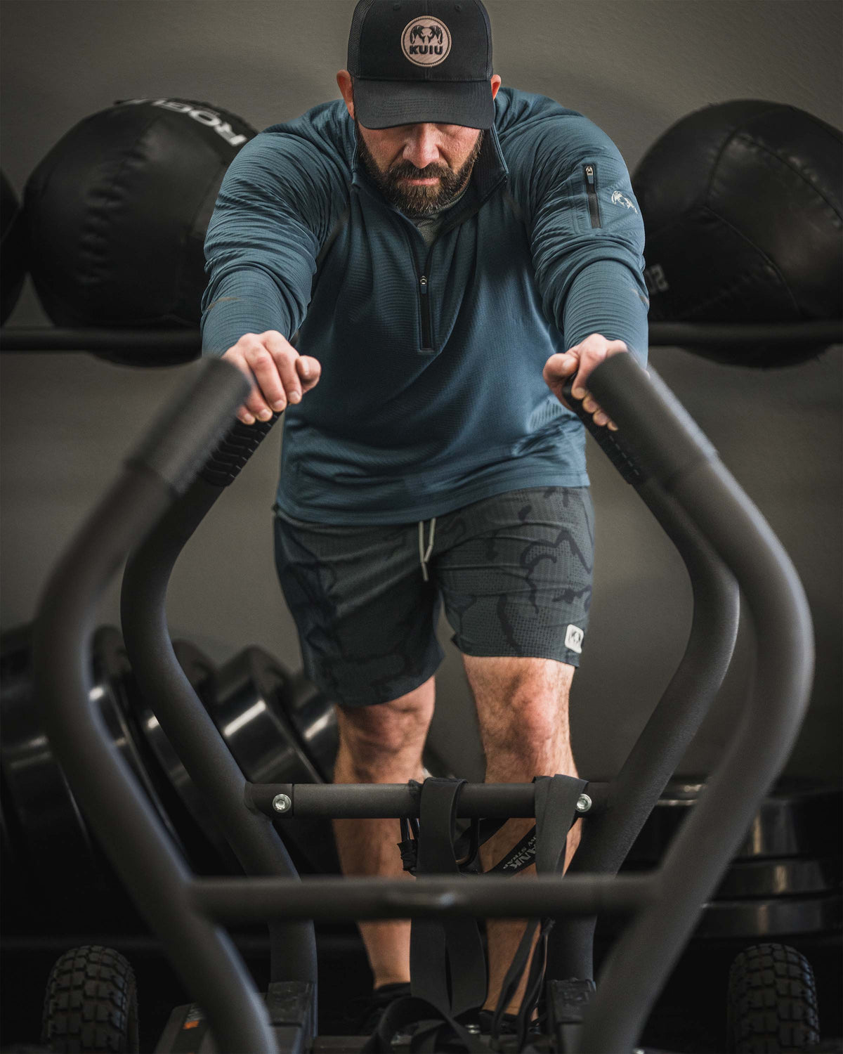 Man training with sled push equipment wearing a black KUIU Circle Patch Hat and KUIU Enduro Trainer Lined Short in Dark Iron Tonal and KUIU Enduro Trainer Zip-T in Flint Blue