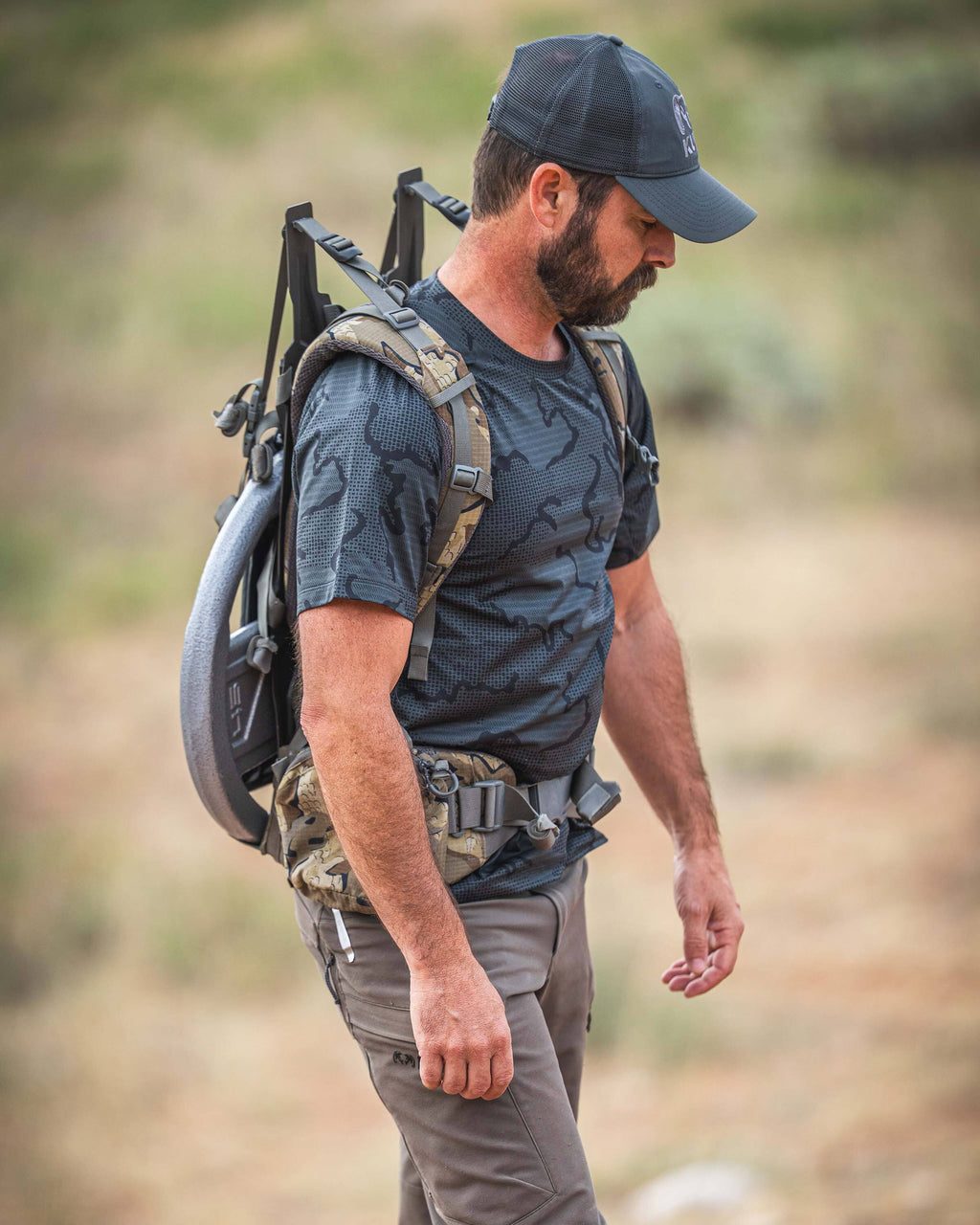 A man wearing a TrailTrainer Short Sleeve Crew in Dark Iron Tonal carrying a large tactical backpack with a weight plate mounted on the back, walking outdoors in a blurred natural setting.
