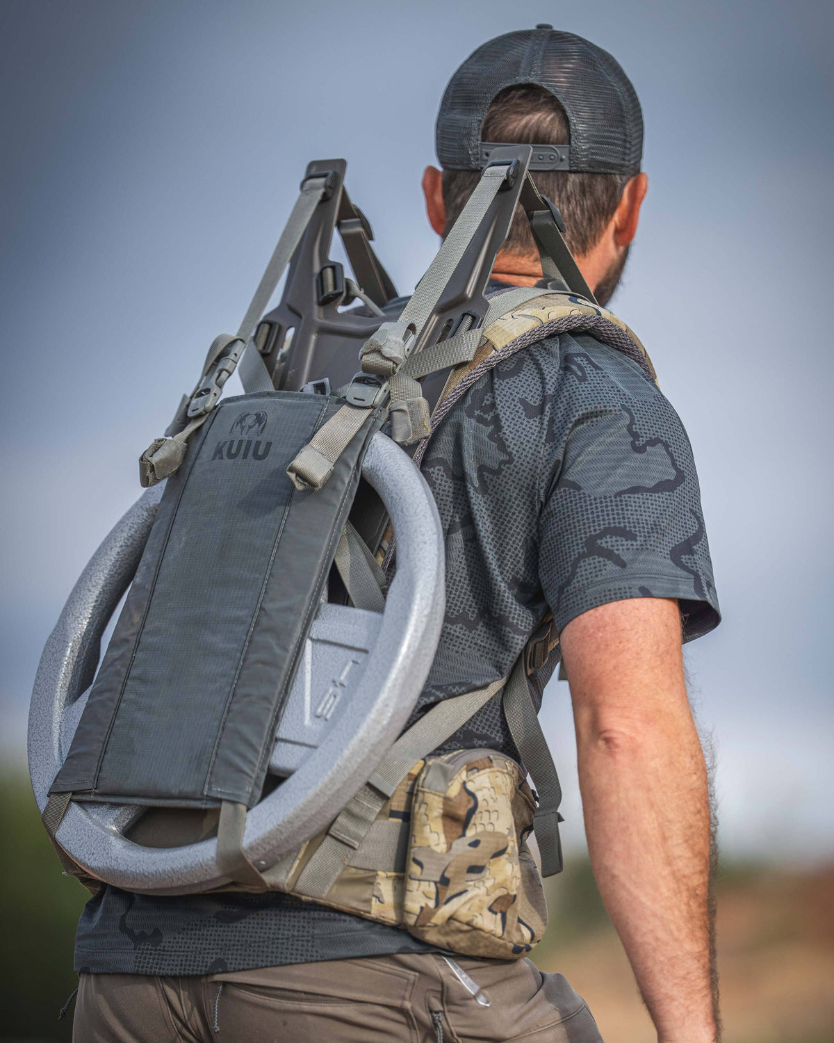 A man in an outdoor setting viewed from behind wearing a TrailTrainer Short Sleeve Crew in Dark Iron Tonal and Valo camouflage suspension and frame with a large weight plate attached.