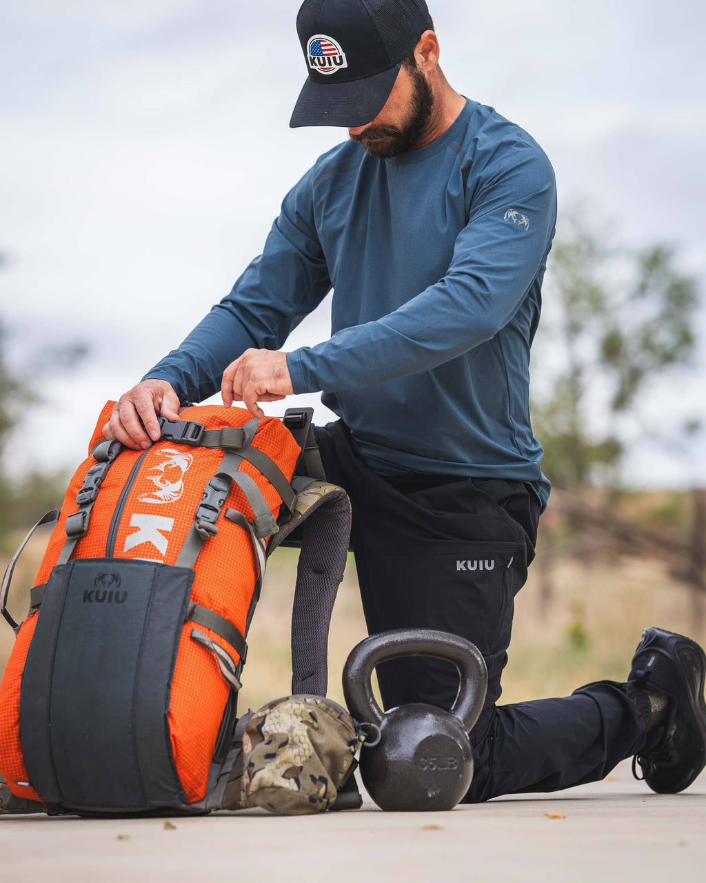 A man wearing TrailTrainer Long Sleeve Crew in Flint Blue kneels next to a kettleball and a KUIU Extreema Bag in Orange.