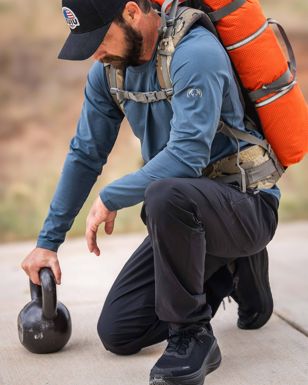 A man wearing TrailTrainer Long Sleeve Crew in Flint Blue kneels next to a kettleball and a KUIU Extreema Bag in Orange strapped on his back.