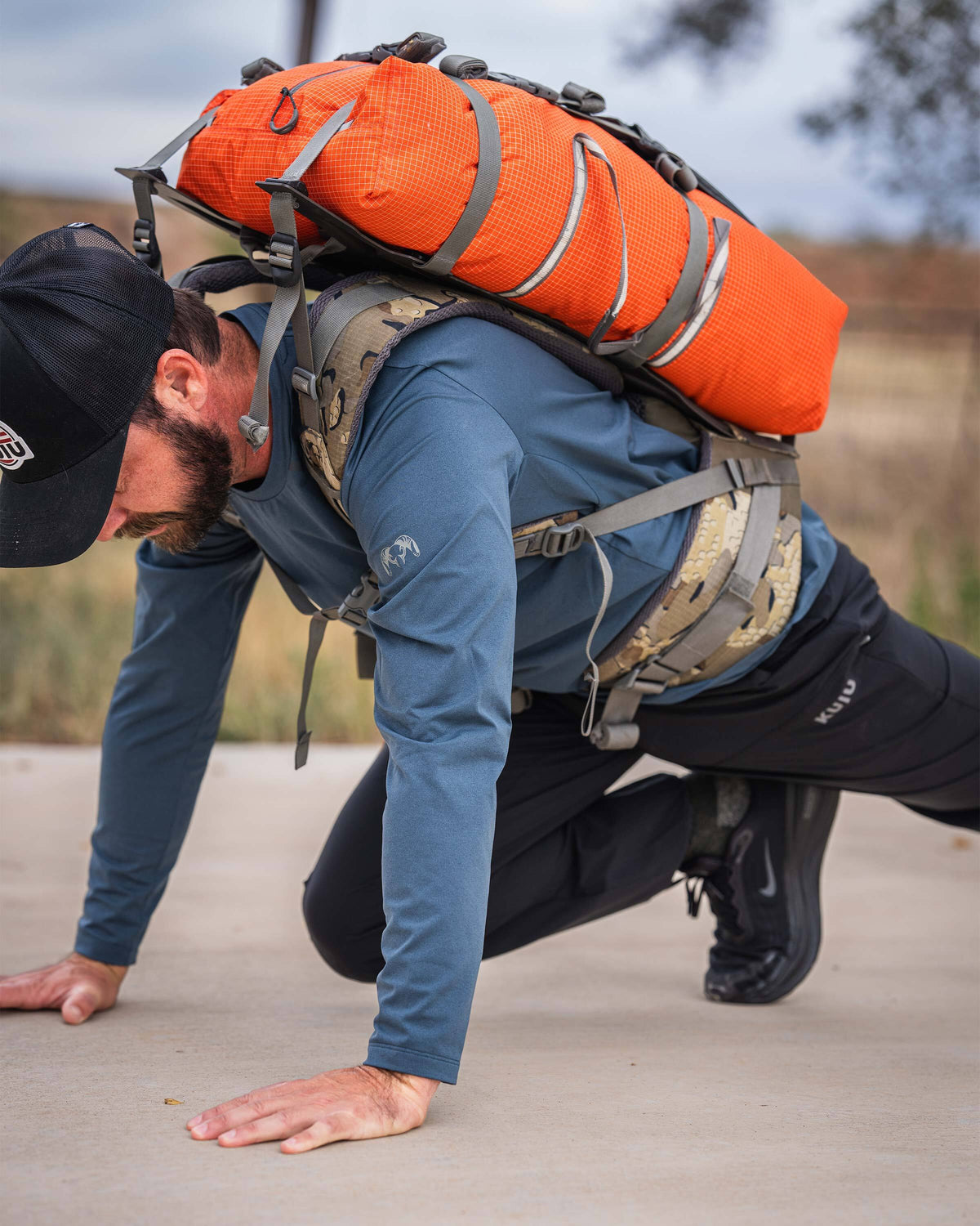 Man in a mountain climbers exercises position carrying a harnessed Extreema Bag in Orange on his back, wearing TrailTrainer Long Sleeve in Flint Blue.