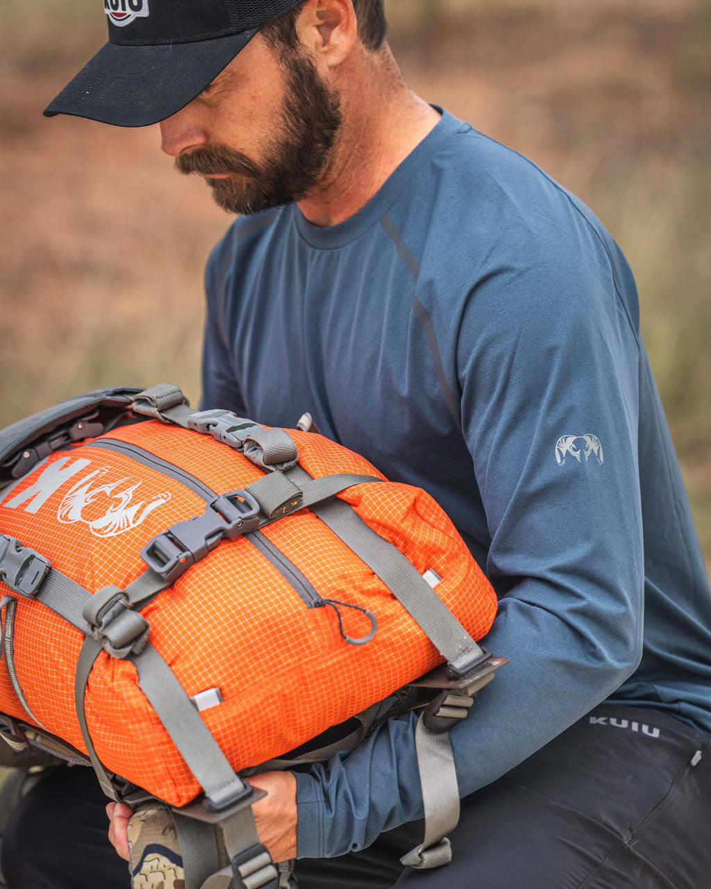 A man holding a harnessed KUIU Extreema Bag in Orange wearing TrailTrainer Long Sleeve in Flint Blue.
