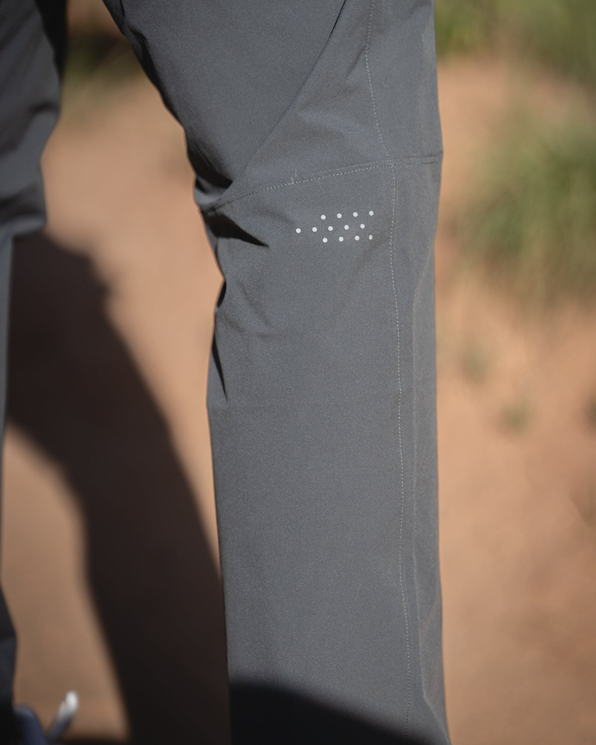 Close-up of TrailTrainer Pant in Black with a small white reflective dot pattern behind knees, shown outdoors on a sandy ground.