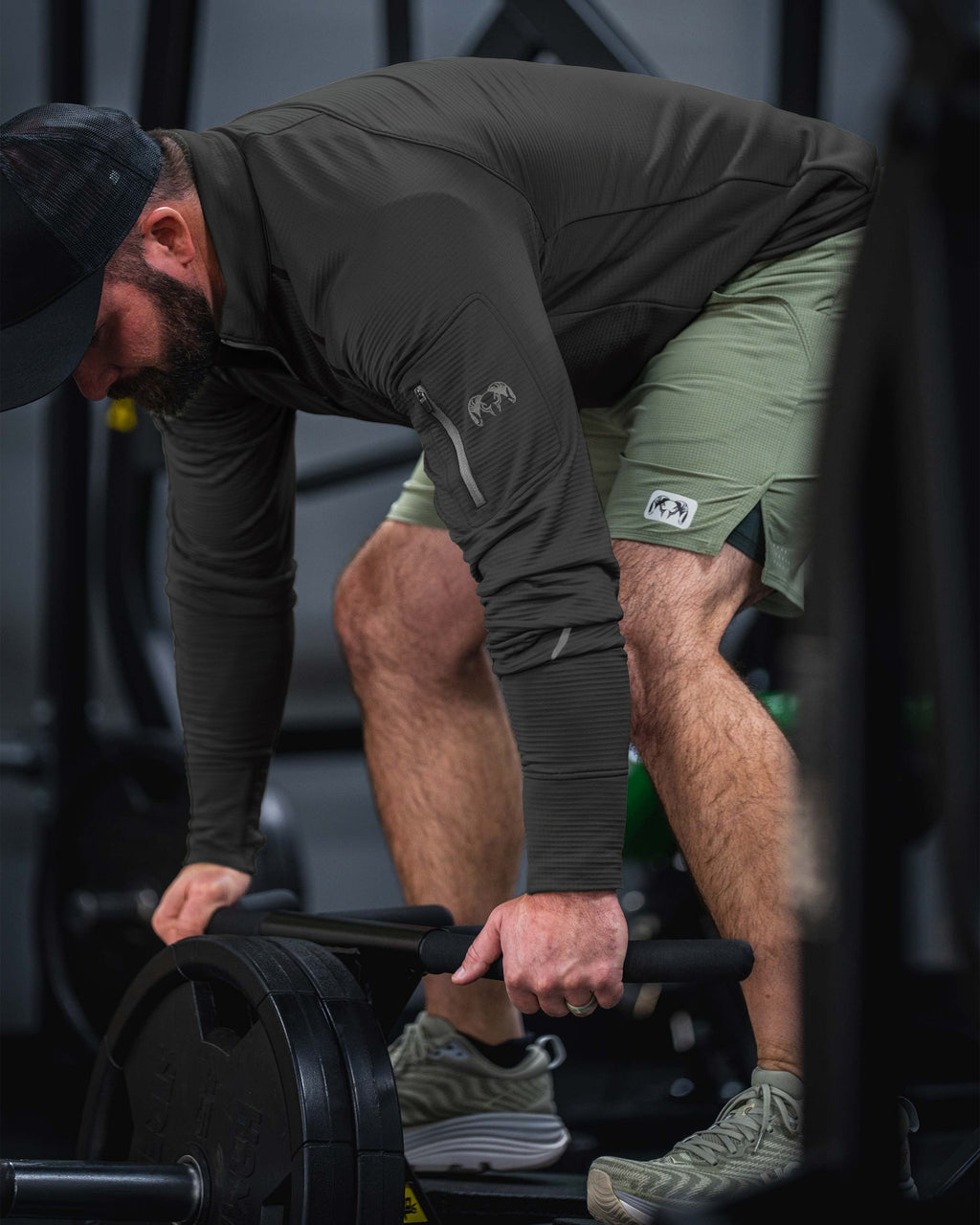 Man wearing KUIU EnduroTrainer Zip-T in Dark Iron Grey and KUIU EnduroTrainer Lined 7" Short in Antler Khaki bending over to lift a heavy barbell