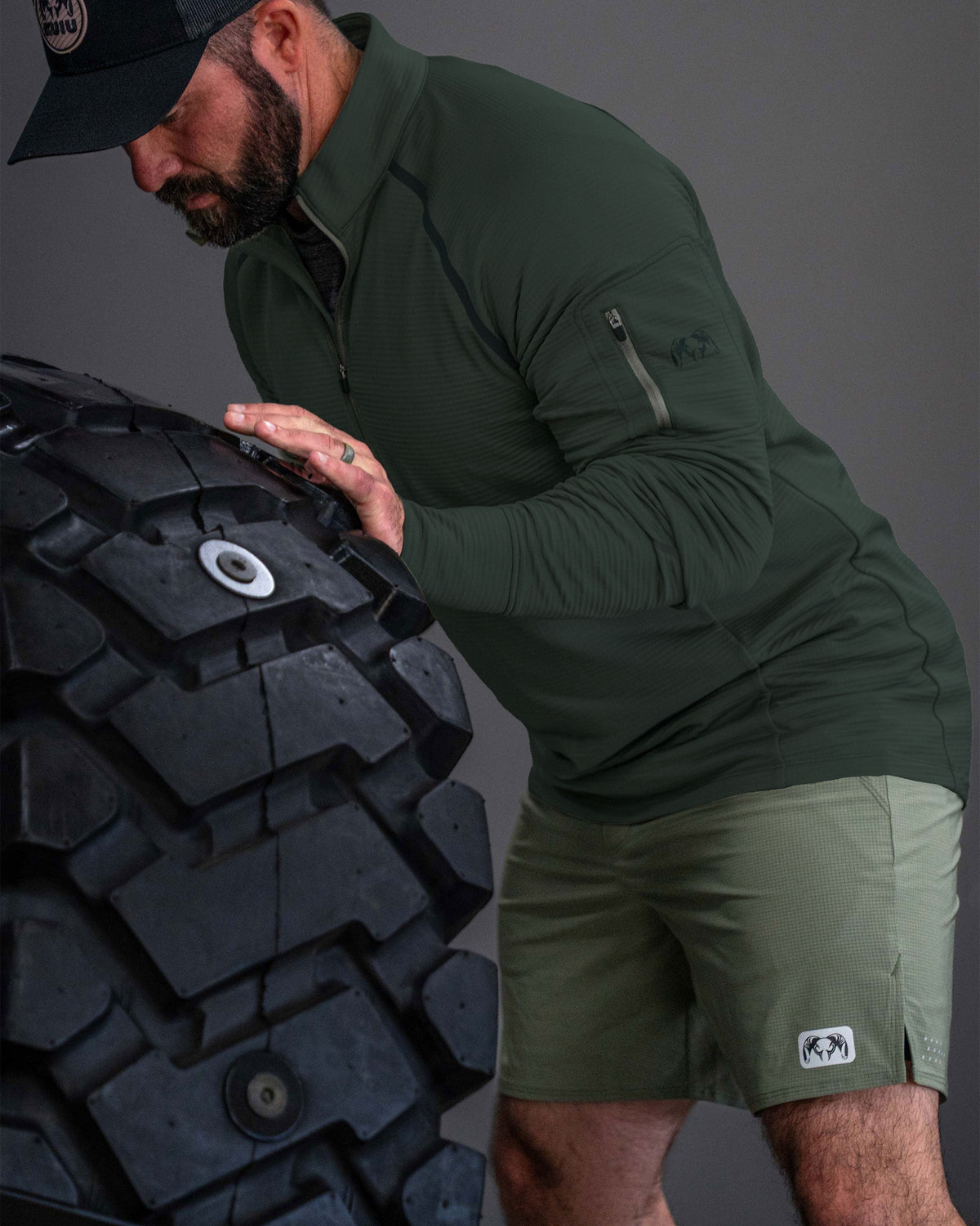 Man leaning forward and pushing an oversized upright rubber tire, performing strength training while wearing KUIU EnduroTrainer Zip-T in Dark Iron Grey and EnduroTrainer 7" Short in Antler Khaki