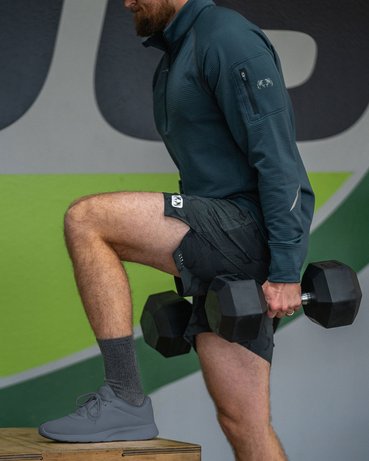 Man performing a step-up exercise on a wooden box while holding two black hexagonal dumbbells wearing KUIU EnduroTrainer 7" Short in Dark Iron Tonal Grey