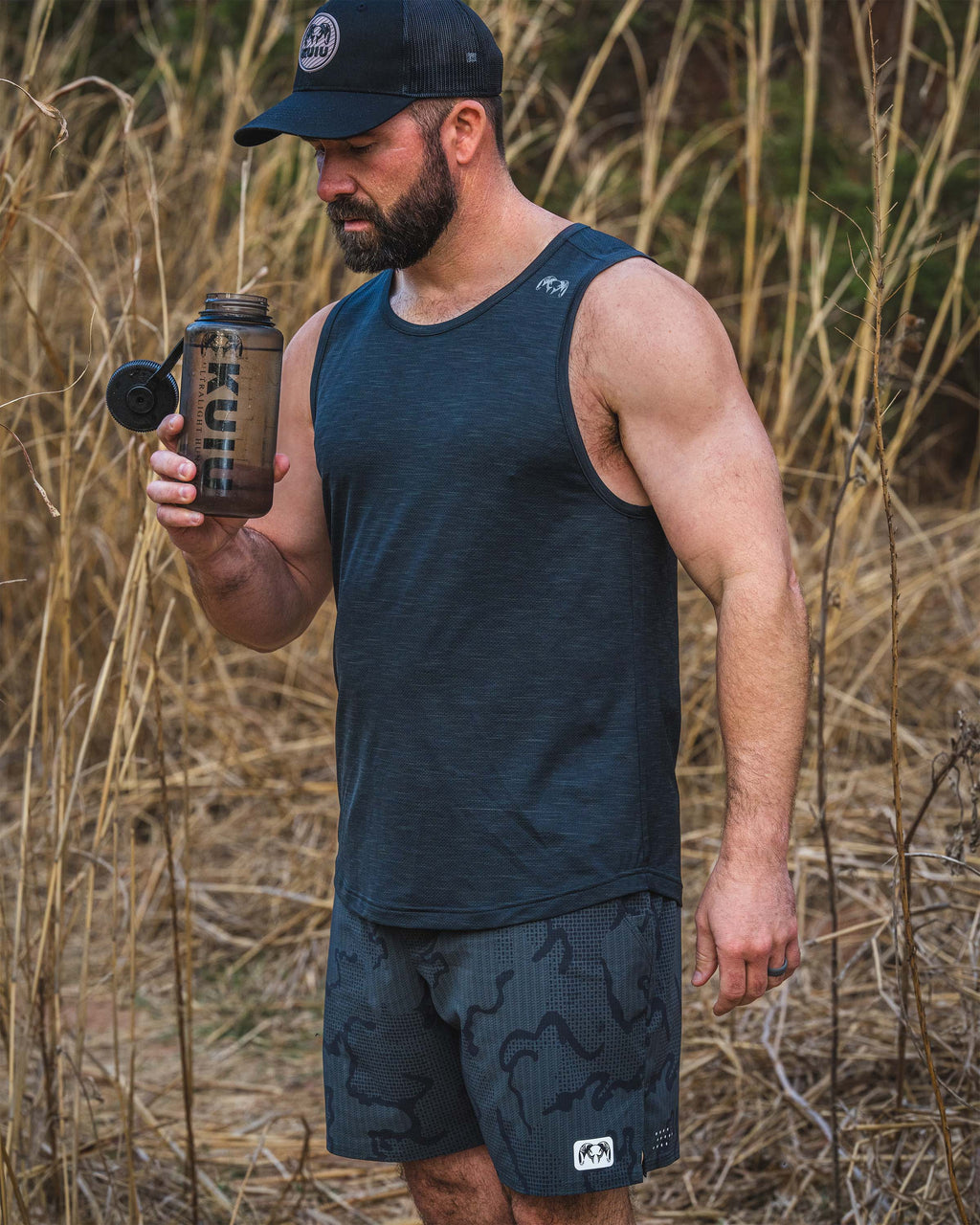 Man wearing an EnduroMesh Tank in Black stands outdoors in tall dry grass, holding and a translucent KUIU 32 oz Wide Mouth Bottle with a flip lid.
