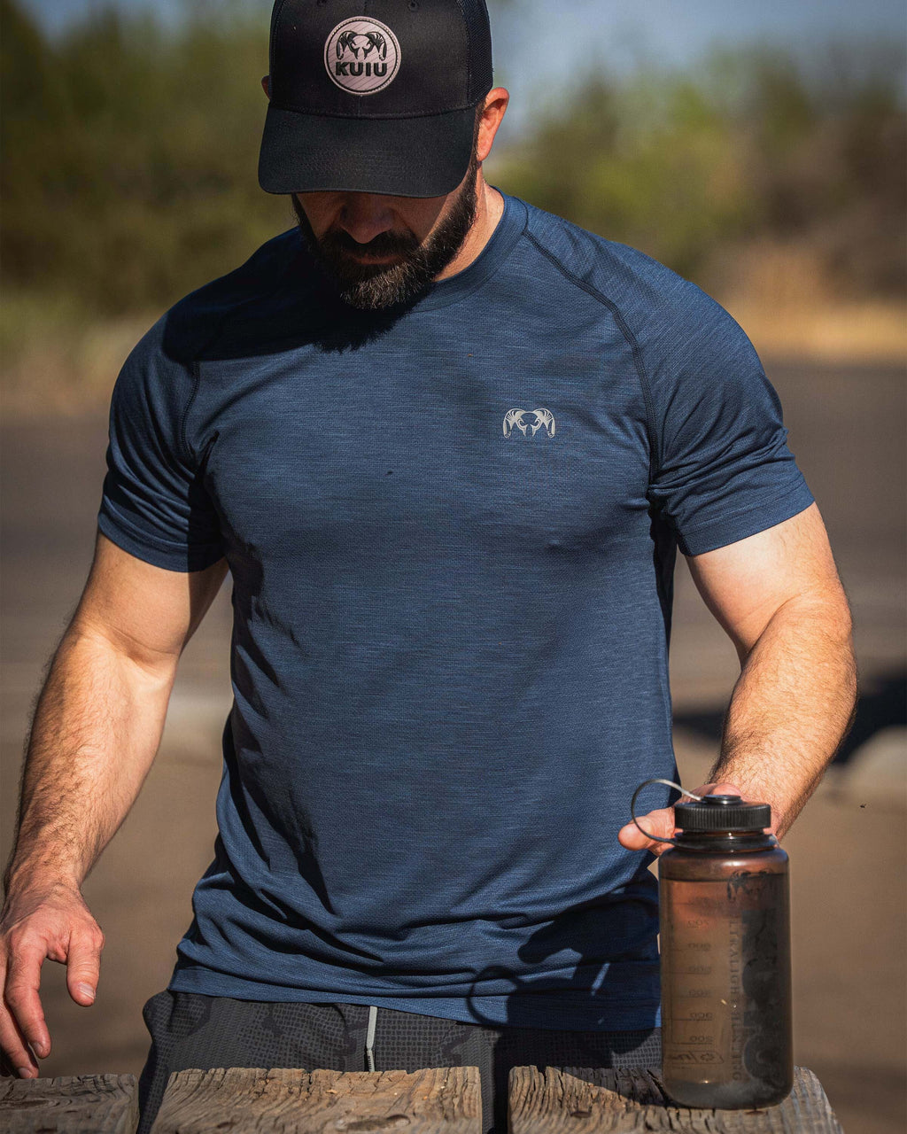 Man wearing EnduroMesh Short Sleeve Crew in Midnight Blue Heather near a wooden surface, holding a translucent KUIU 32 oz Wide Mouth Bottle.
