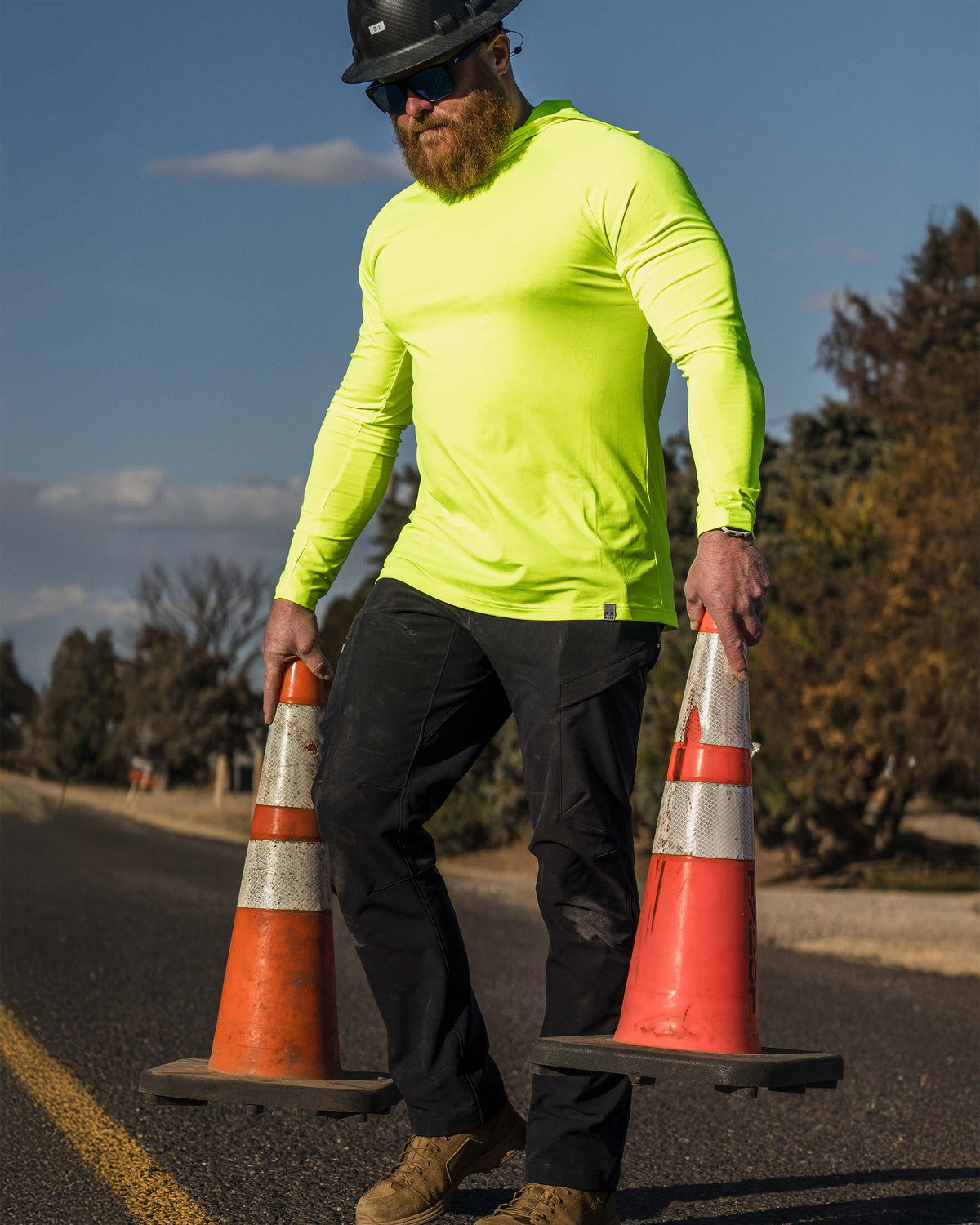 Man is wearing Workgrade Sun Hoodie in Highlight color, a bright, high-visibility long‑sleeve shirt in a neon yellow‑green color, holding two orange traffic cones one in each hand. 