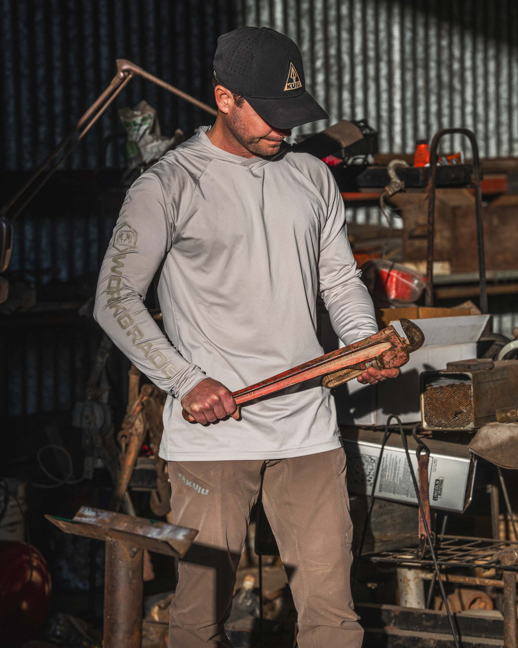 Man in a workshop, holding a large rustic wrench, wearing Workgrade Long Sleeve Sun Hoodie in Haze Grey. 