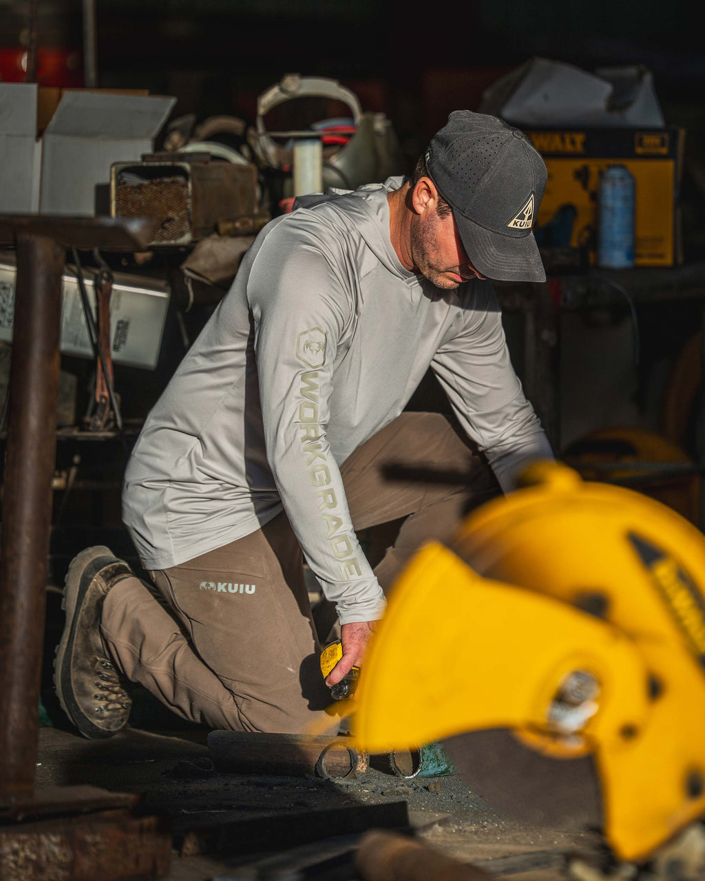 Man kneeling in a workshop using a yellow power tool to cut metal, wearing Workgrade Long Sleeve Sun Hoodie in Haze Grey