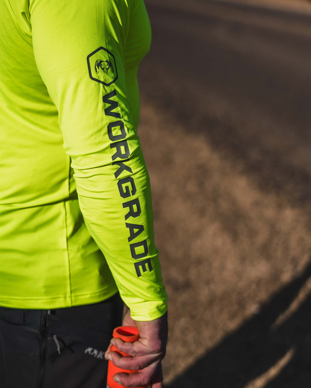 Close-up of a man wearing Workgrade Long Sleeve Sun Hoodie in neon yellow‑green Highlight color with “WORKGRADE” printed down the sleeve, holding an orange traffic cone at their side.
