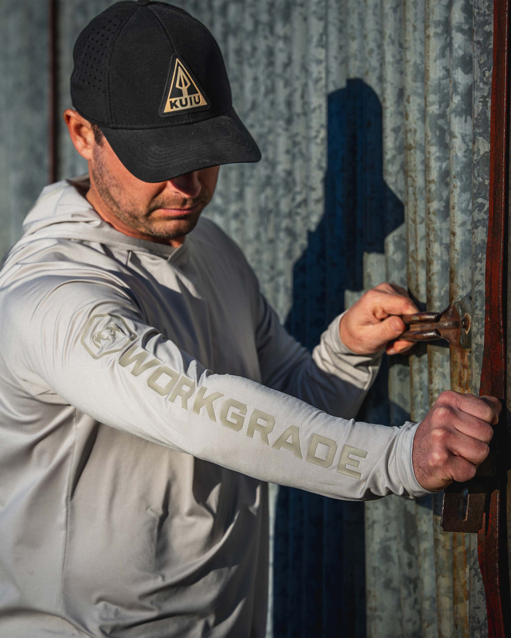 Man closing a large rustic metal door, wearing Workgrade Long Sleeve Sun Hoodie in Haze Grey with “WORKGRADE” printed down the right sleeve.
