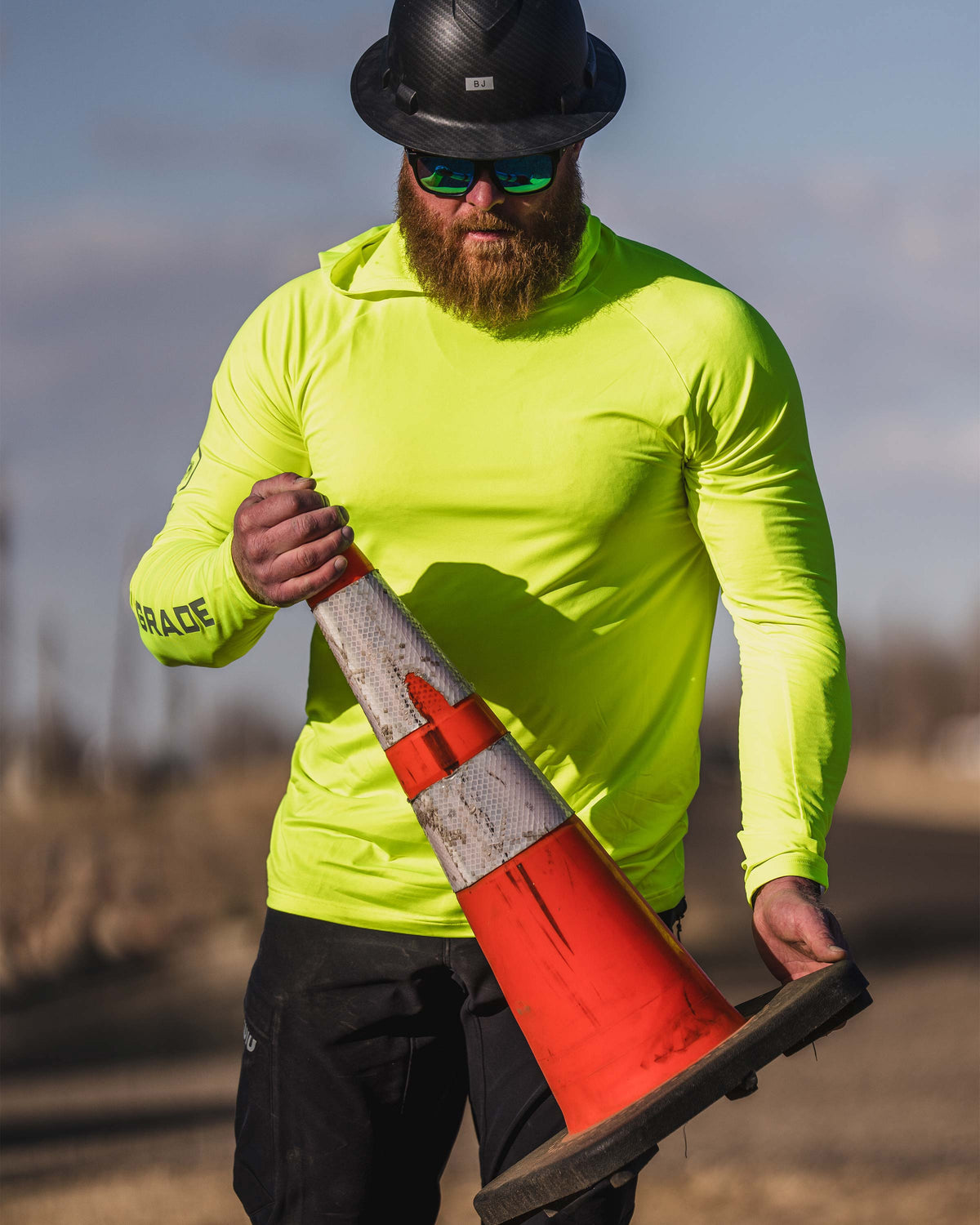 Man in an outdoor environment, wearing Workgrade Long Sleeve Sun Hoodie in neon yellow‑green Highlight color. Both hands are holding an orange traffic cone.