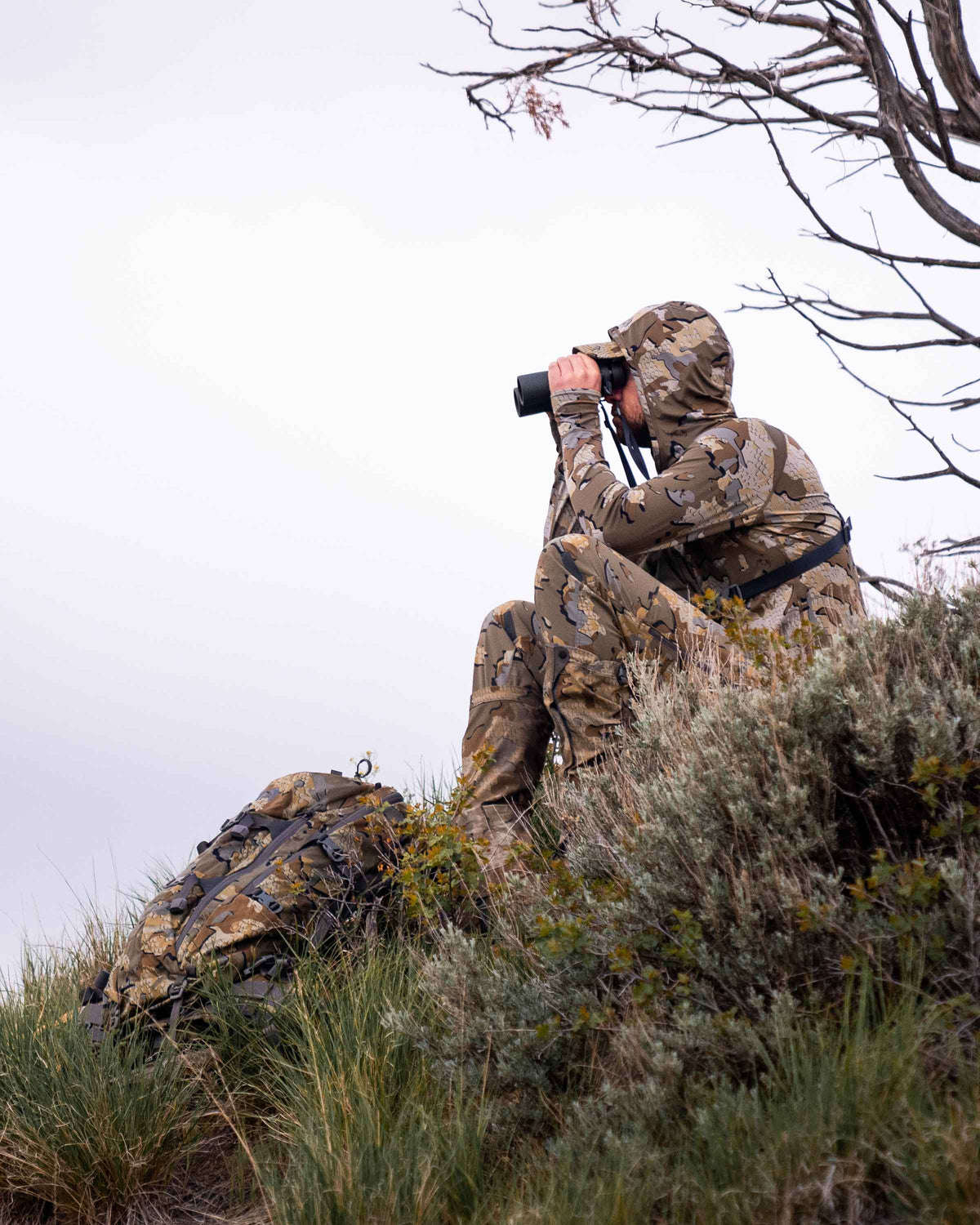 Hunter sitting down, glassing on a grassy hillside, wearing Gila Long Sleeve Hoodie in Valo Camouflage