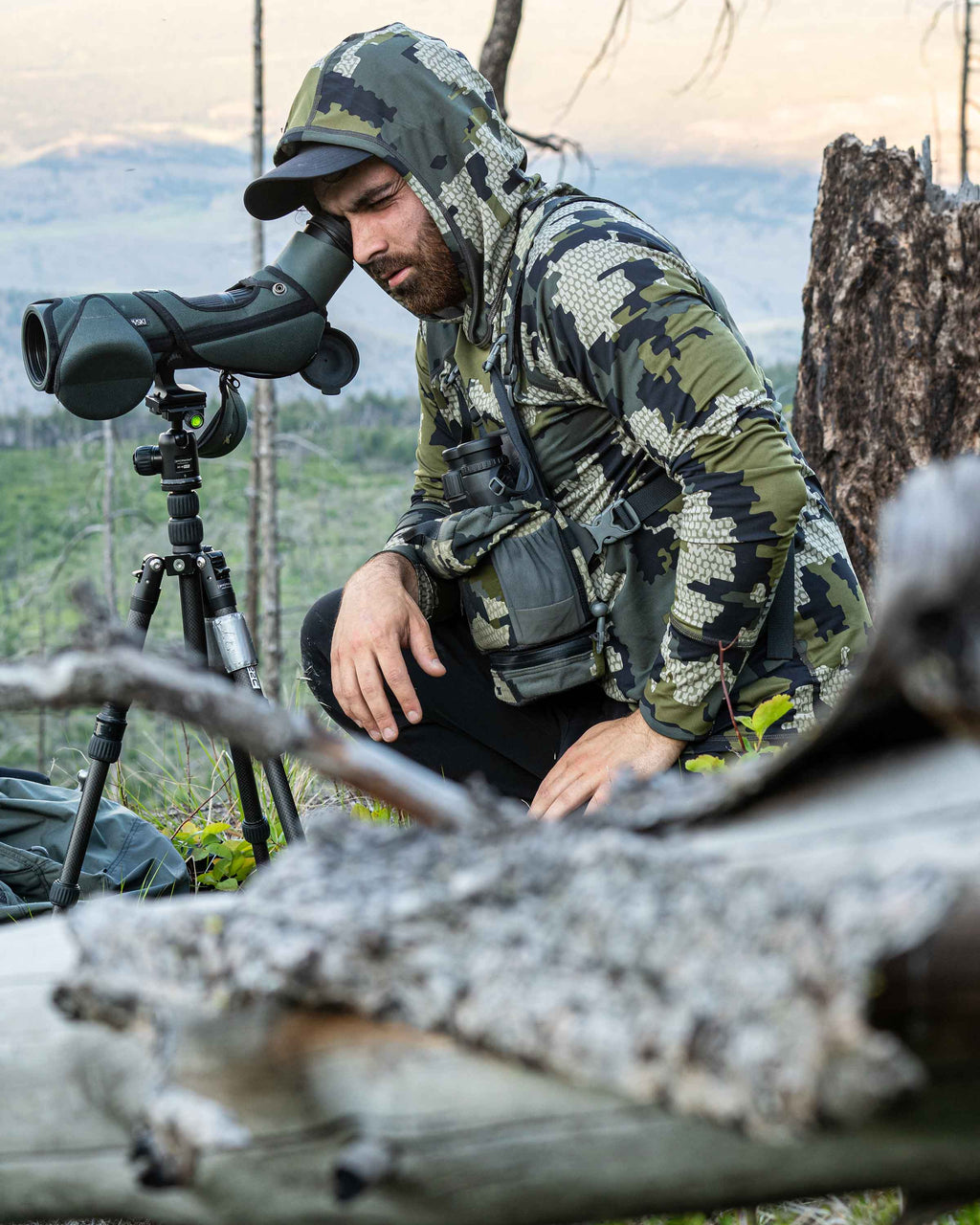 Hunter kneeled down by a log looking into a spotting scope, wearing Gila Long Sleeve Hoodie in Verde Camouflage