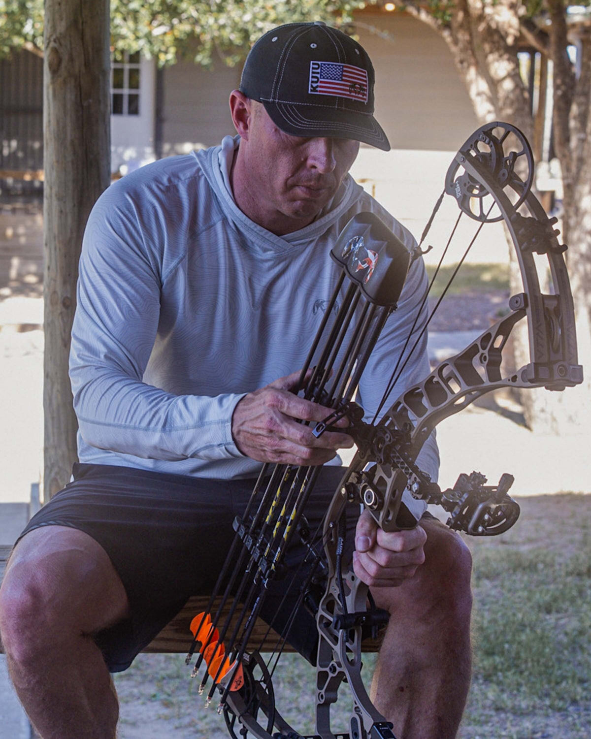 Man sitting in shade, holding compound bow, wearing Gila LS Hoodie in Skyline White Topo Print
