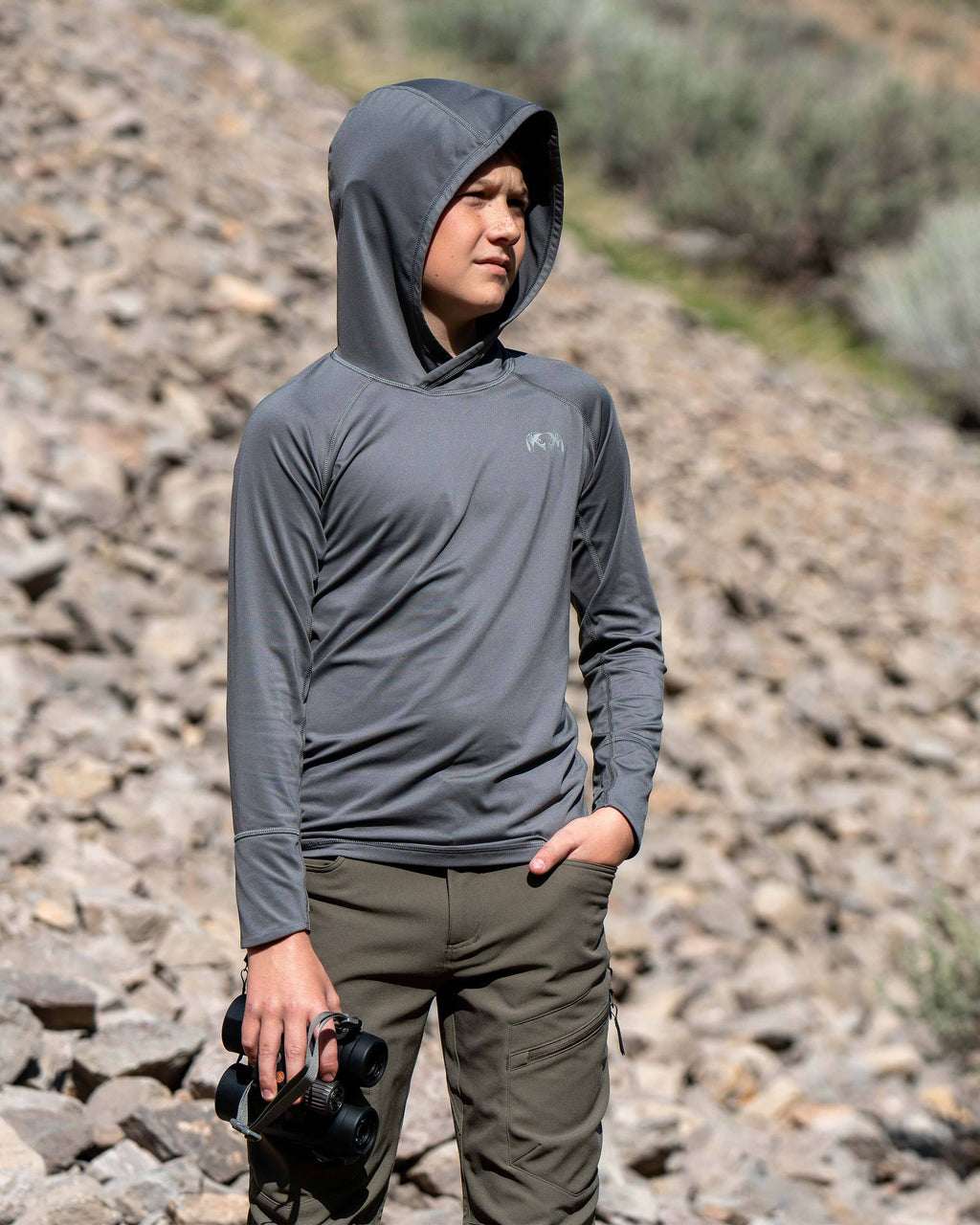 Youth wearing Youth Gila LS Hoodie in Stone Grey, holding a binocular in his right hand in a rocky terrain environment