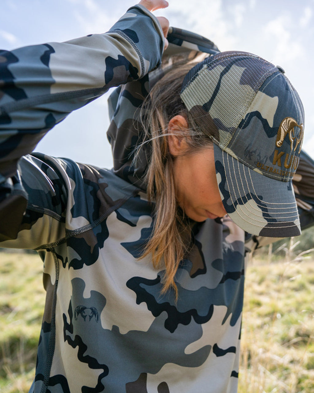Female hunter putting on hood wearing Women's Gila LS Hoodie in Vias Camouflage