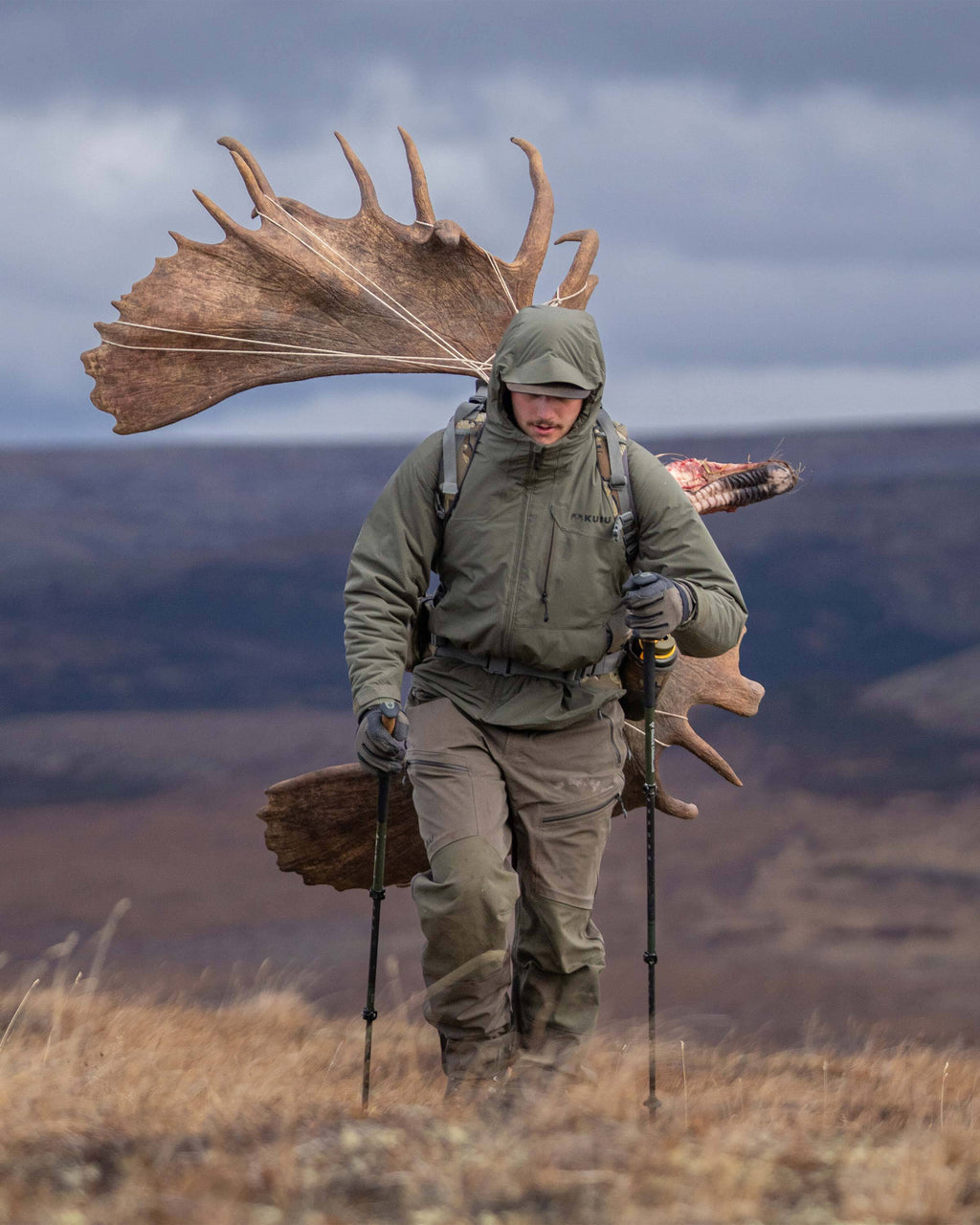 Hunter trekking up a hill carrying a large, palmated moose antlers on his back, wearing Mountain PRO Hooded Jacket in Ash Brown